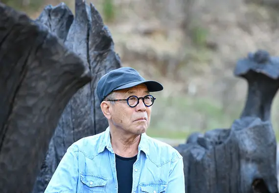 A man in a blue shirt and hat looks off camera, in front of a rock or tree formation.