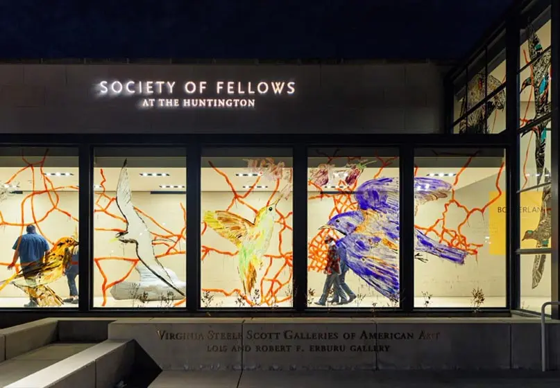 The painted glass panels of the Virginia Steele Scott Galleries of American Art are lit up at night while a couple of people walk behind the painted glass. Projected on the roof, outside, is "Society of Fellows at The Huntington"