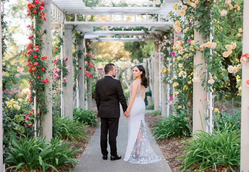 A bride and groom stand holding hands under an arbor of colorful climbing roses.