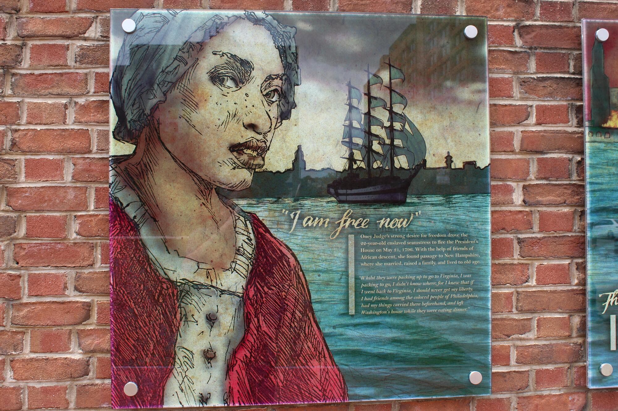 Image of a museum sign with an artist's rendition of Ona Judge, an enslaved woman who escaped from the home of her enslavers, George and Martha Washington, in 1796. The words "I am free now" are visible beneath the image of a ship in Philadelphia's harbor. The sign is mounted on a red brick wall.