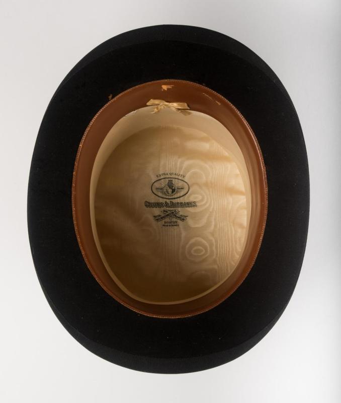 Underside view into interior of black beaver-skin top hat, with label that reads Collins and Fairbanks Co. sitting on plain surface.