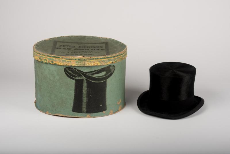 Black beaver-skin top hat placed beside a light turquoise hat box; the side of the box has pasted print of an upside down black top hat ; both hat and box are placed against a plain, white background