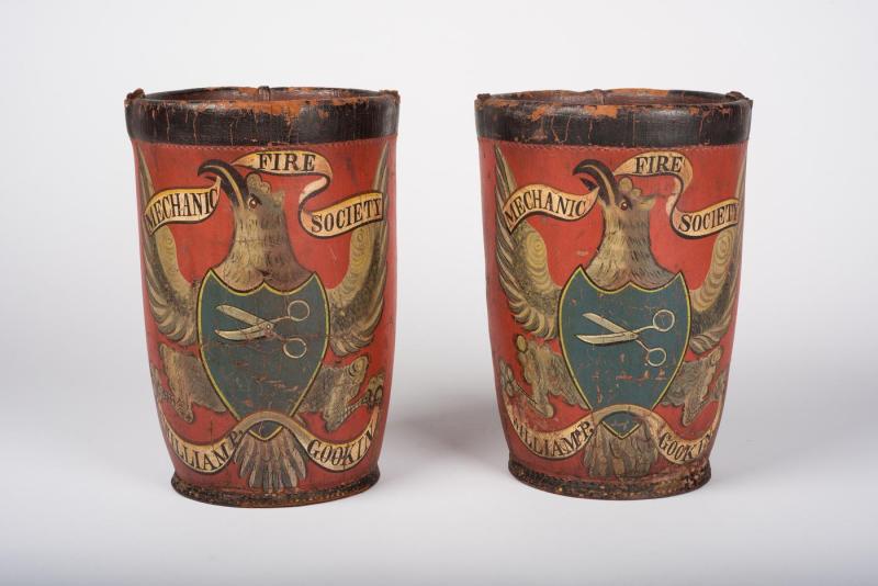 Two red leather buckets decorated with eagles bearing blue shields with scissors. Banners in their beaks read "Mechanic Fire Society"; banners in their talons read "William P. Gookin."