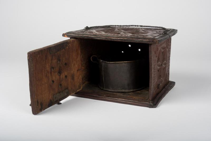 Brown wooden box foot-warmer with perforated star-shaped designs on its sides and top with a panel open revealing its cylindrical metal heating element used for holding hot coals.