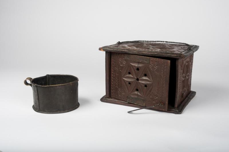 Brown wooden box foot-warmer with perforated star-shaped designs carved on its sides and top placed beside its cylindrical metal heating container used to hold hot coals.