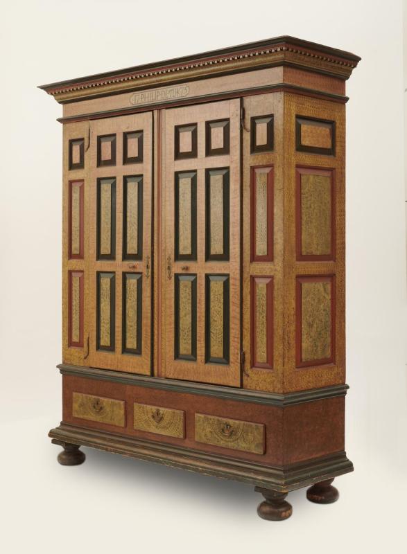 Large wooden wardrobe with panels, each with a border of red, green or black and painted decoration simulating the look of wood grain; drawers across bottom and cornice at top.