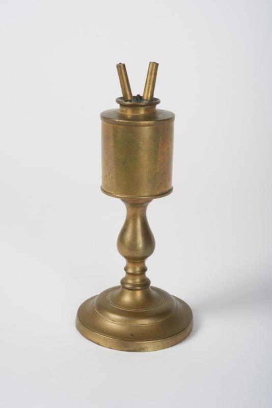 Brass lamp that resembles a large candlestick with a reservoir and two wick holders at the top in the shape of a V.