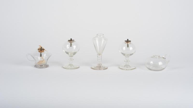 Five glass toy lamps in a variety of shapes, three with feet and bulbous or V-shaped reservoirs, another in a shape resembling a teacup, and another in the shape of a oval sphere. 