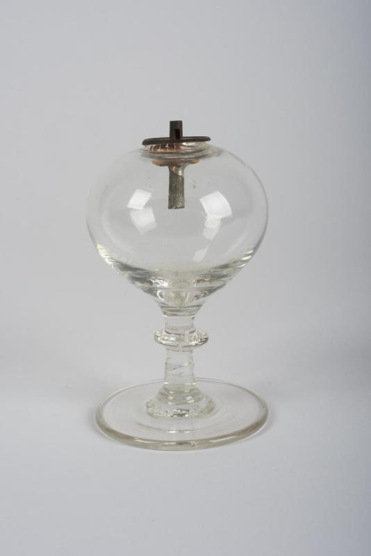 A clear glass toy lamp that was blown by hand; the lamp has an empty round reservoir for kerosene and a tin wick holder and collar at top.