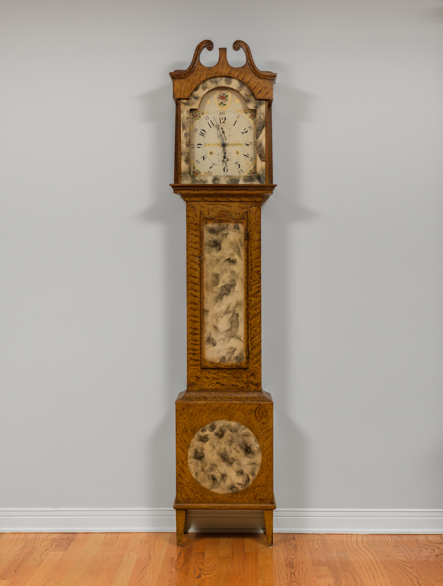 Tall grandfather-style clock in a wooden case painted to resemble different types of wood grains. 