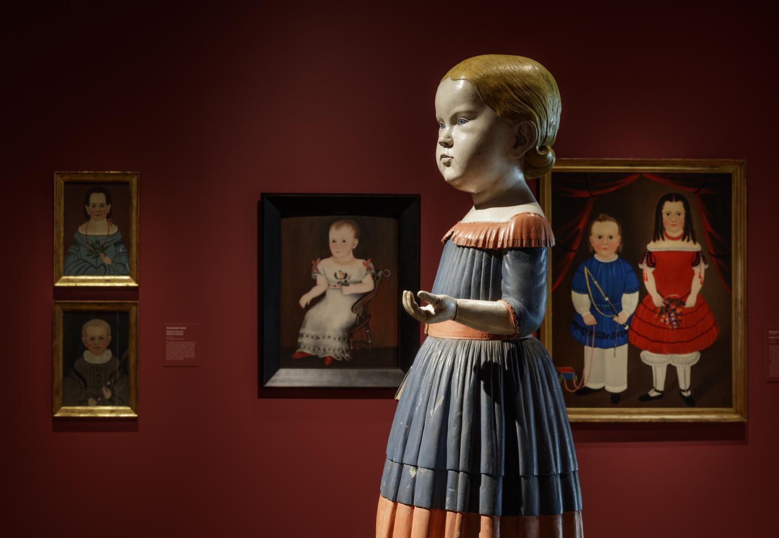 Image of portraits of children displayed on a wall with a sculpture of a young girl placed in front of them.
