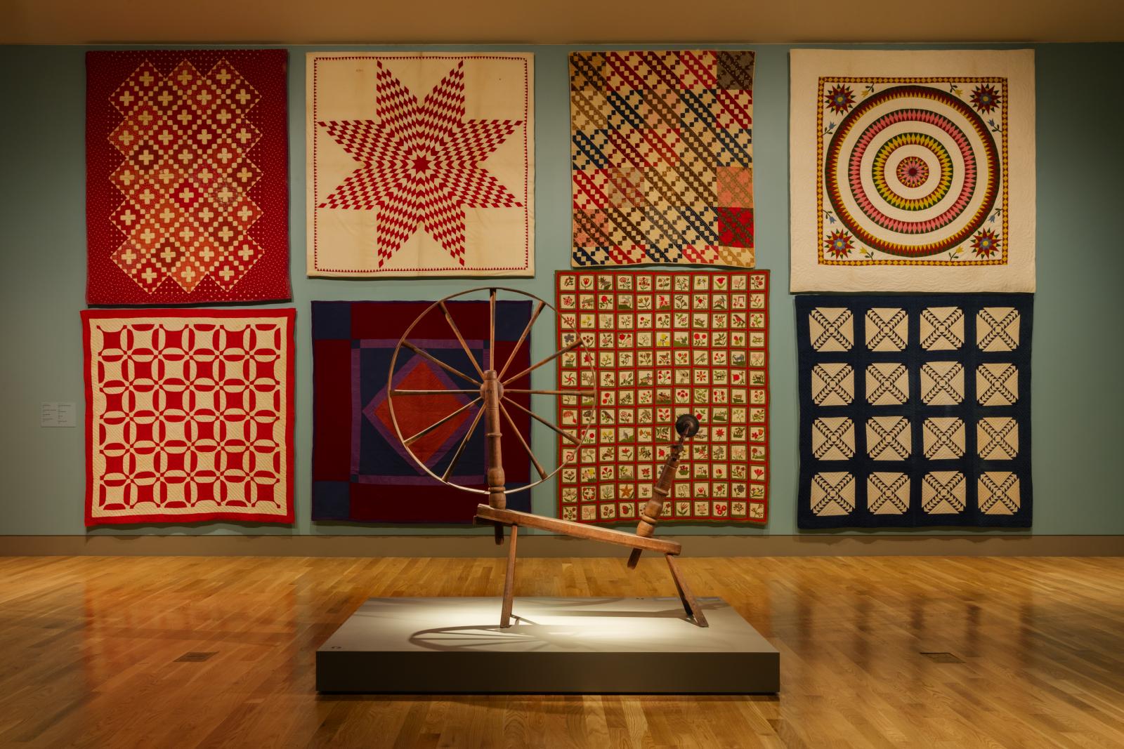 View of eight quilts displayed in a museum gallery with a spinning wheel placed in the center of the room.
