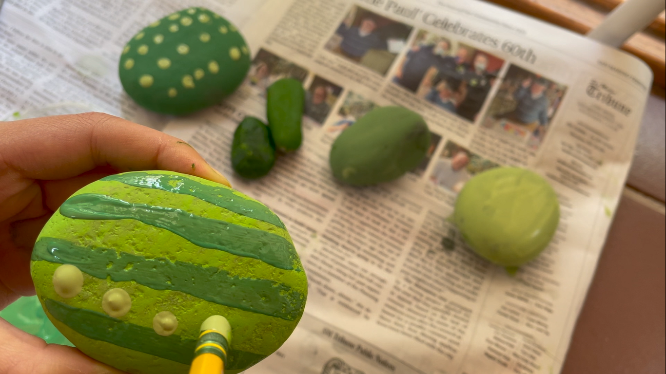 One small circular rock is being painted light green with dark green stripes and dots while other green-painted rocks are blurred in the background.