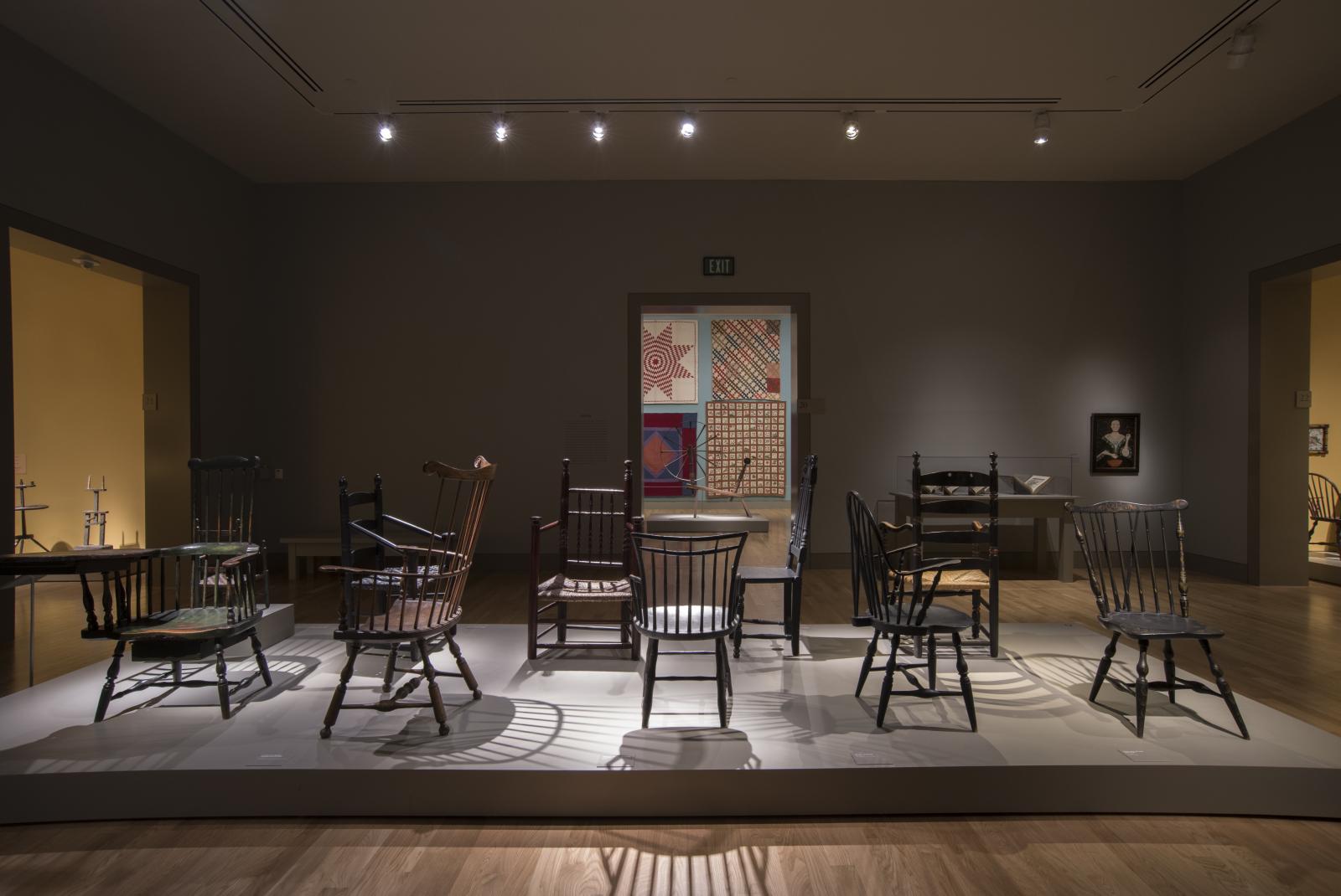 A variety of wooden early American chairs displayed in the center of a dimly lit museum gallery.
