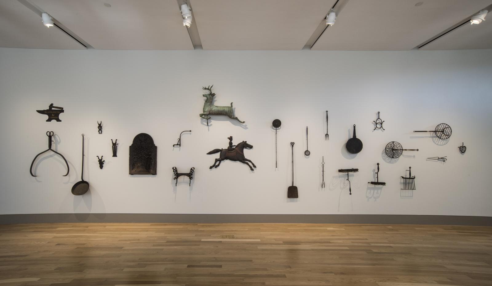 A long gallery wall displaying a variety of early American metal tools of different shapes and sizes.