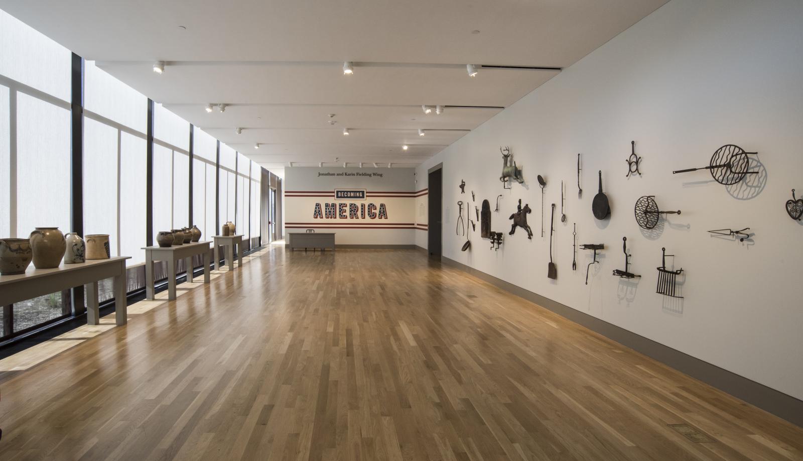 Entrance to the Becoming America exhibition with ceramic jars on tables displayed along one windowed wall and metal tools hanging on another wall.