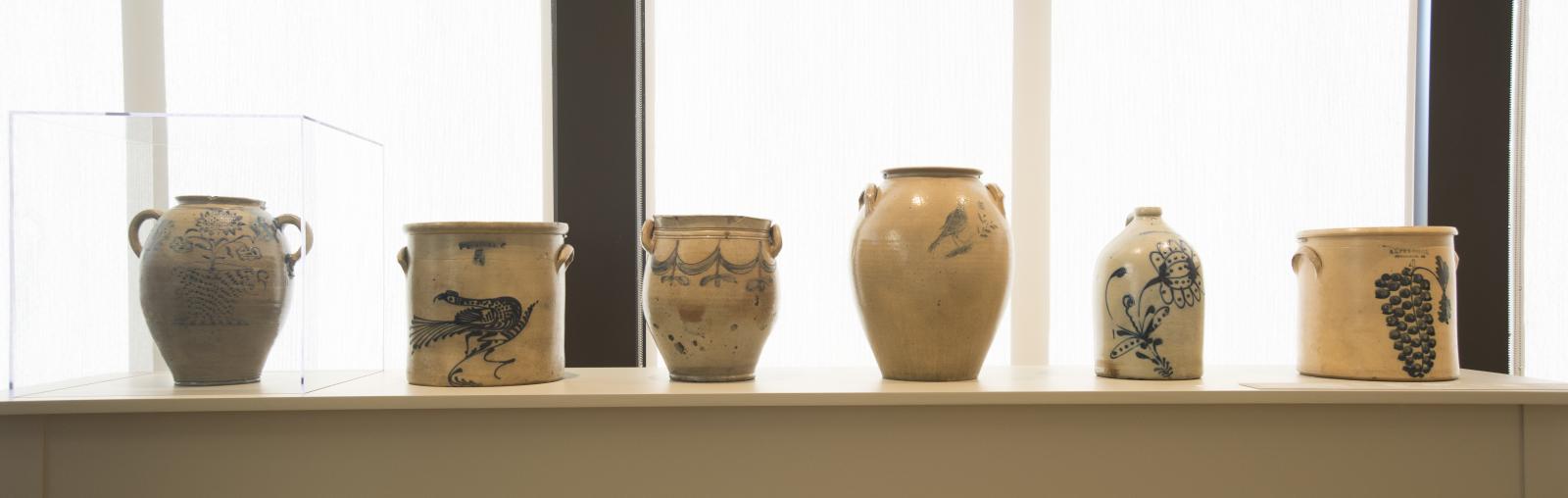Six large ceramic jars with different blue painted details are displayed on a long table in front of a windowed wall.