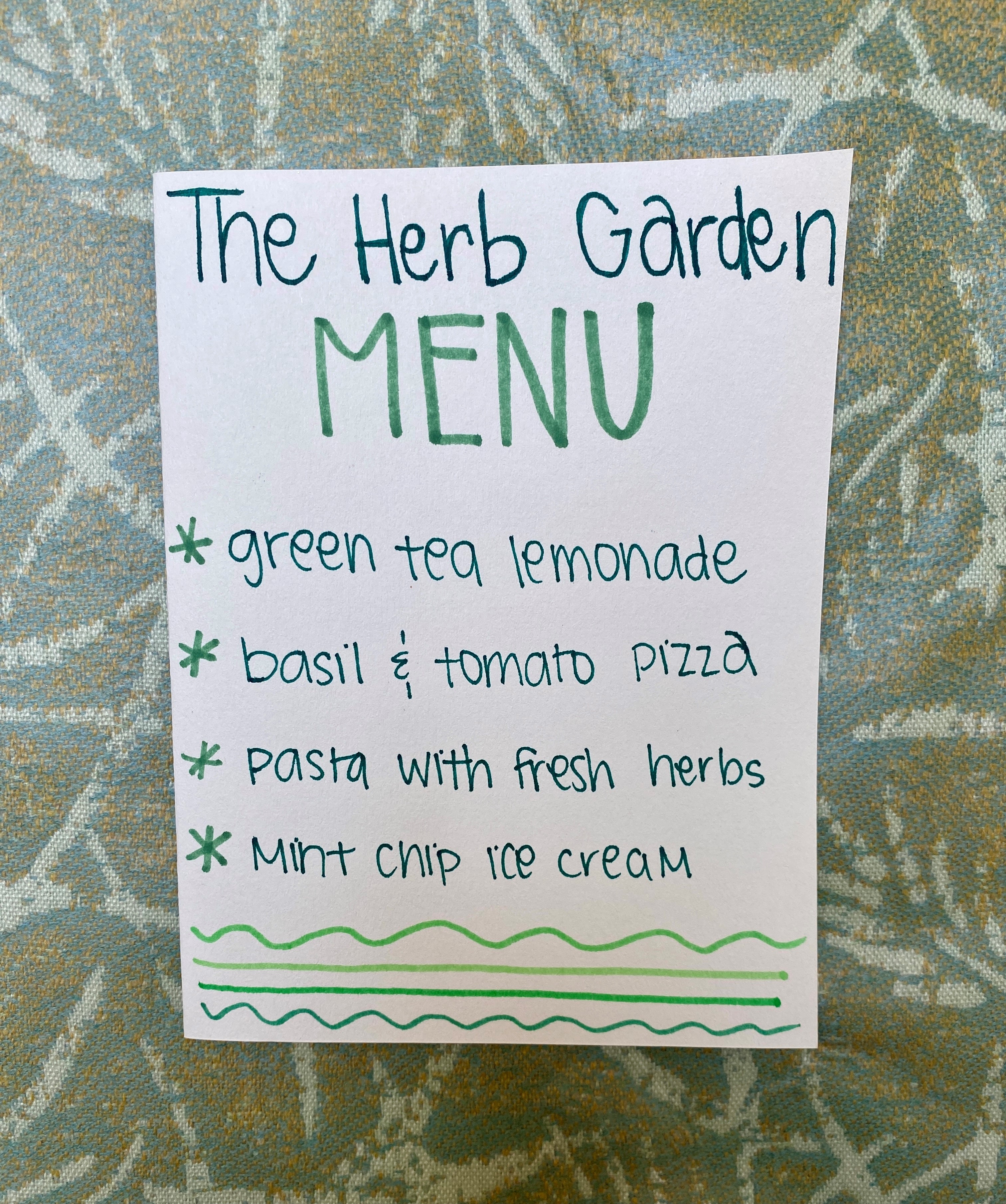 A white piece of paper with handwritten text stating “The Herb Garden Menu, green tea lemonade, basil and tomato pizza, pasta with fresh herbs, and mint chip ice cream” against a green background.