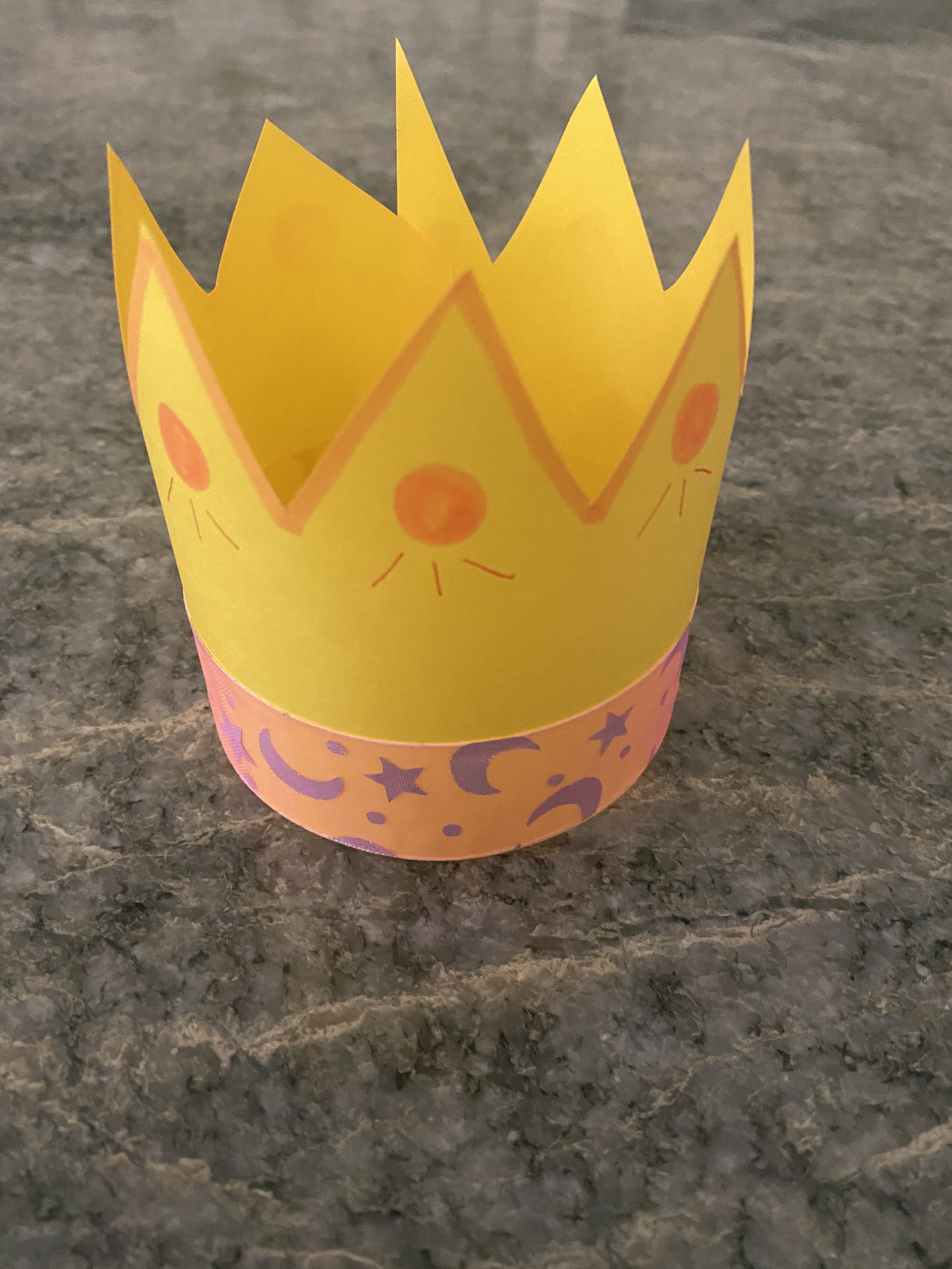 A small yellow crown with a pink and purple star ribbon on the base and a decoration near the triangle points on the top. 