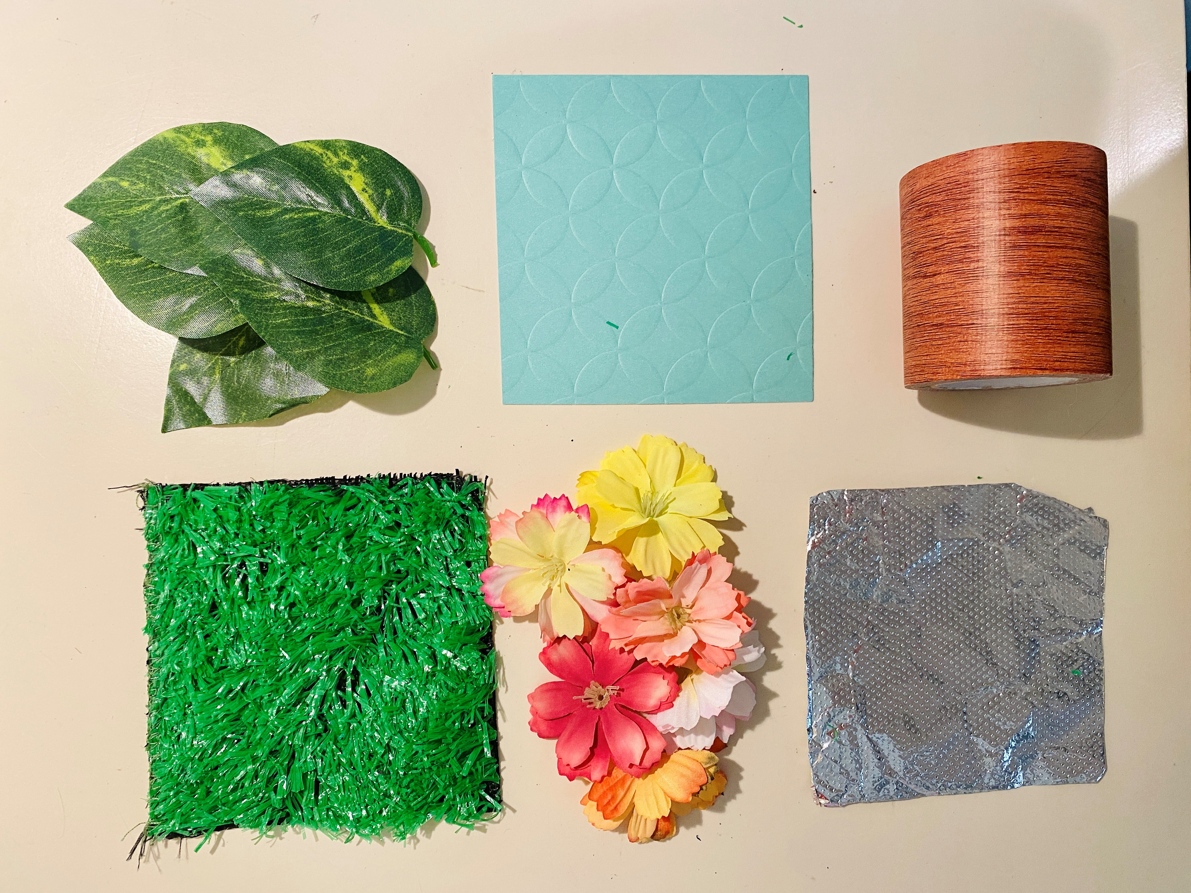  Two rows of materials including green leaves, blue paper, brown tape on the top row and fake green grass, orange and yellow flowers, and aluminum foil on the bottom row.