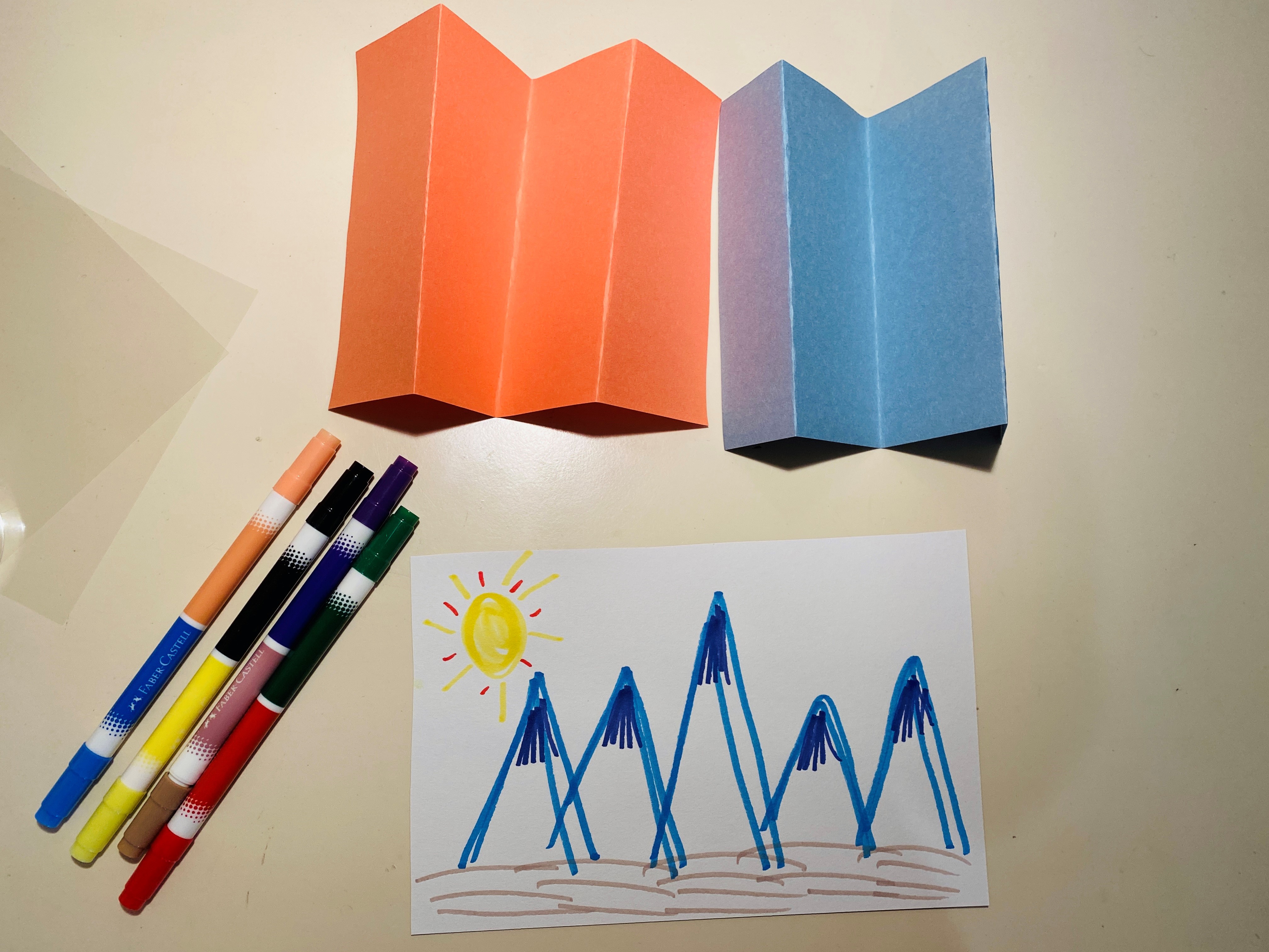 Two folded pieces of paper, one orange and one light blue, four markers and a piece of paper drawn with blue mountains and a yellow sun.