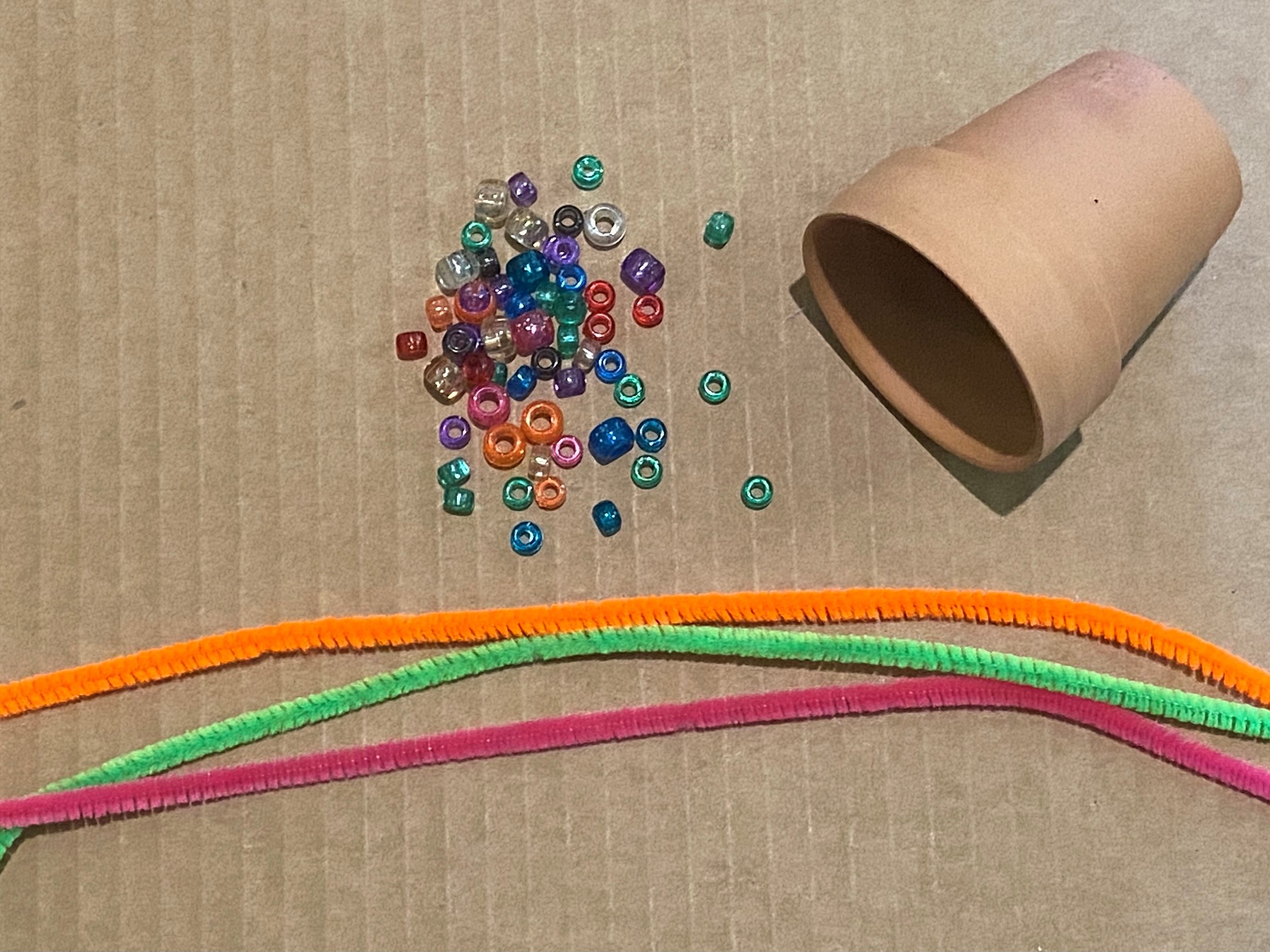 Multi-colored beads, an orange, green, and pink pipe cleaner, and a small brown pot arranged on a brown board.