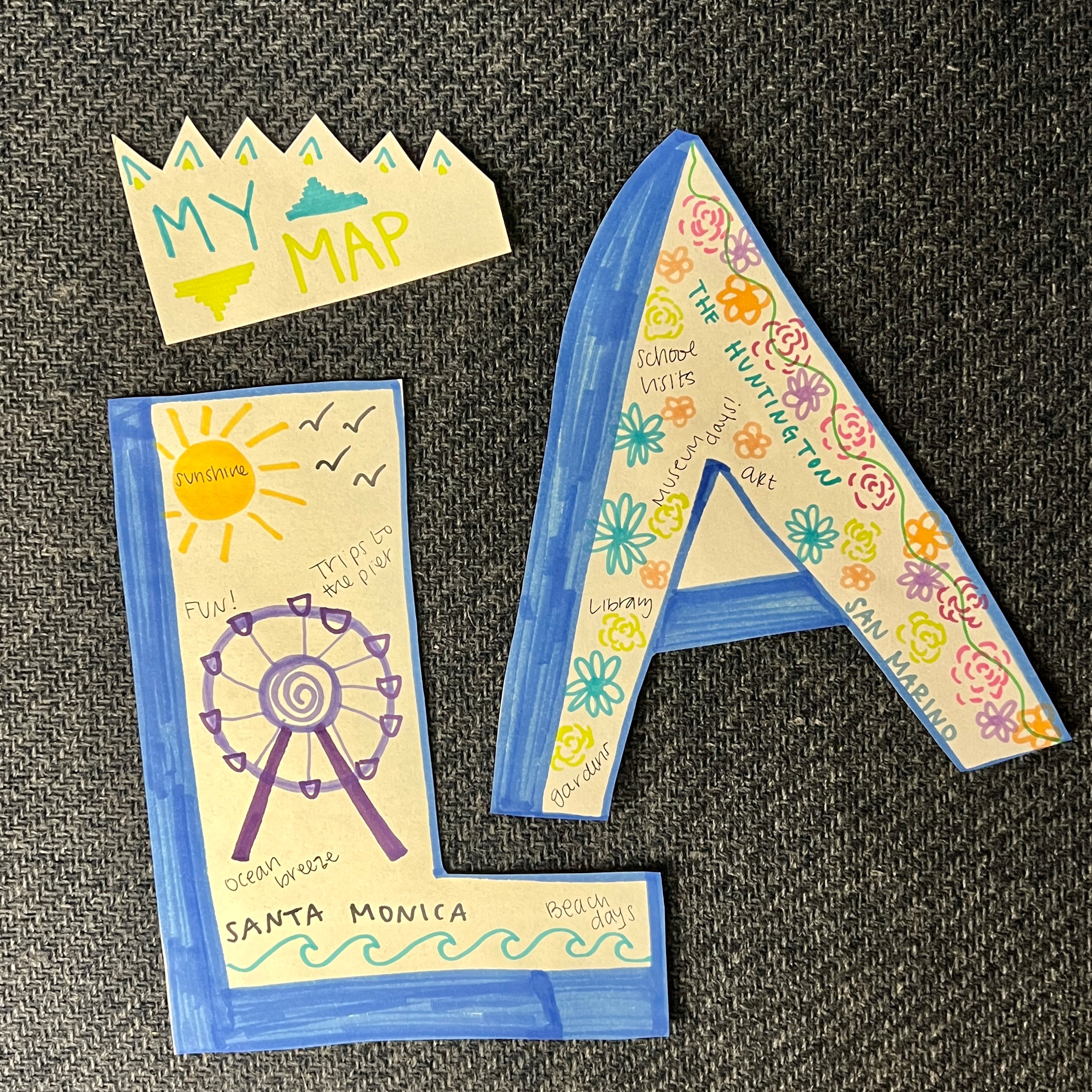 A cutout "L" with a Ferris Wheel and sun drawn inside, a cutout "A" with flowers drawn inside, and a cutout shape that says "My Map" inside. 