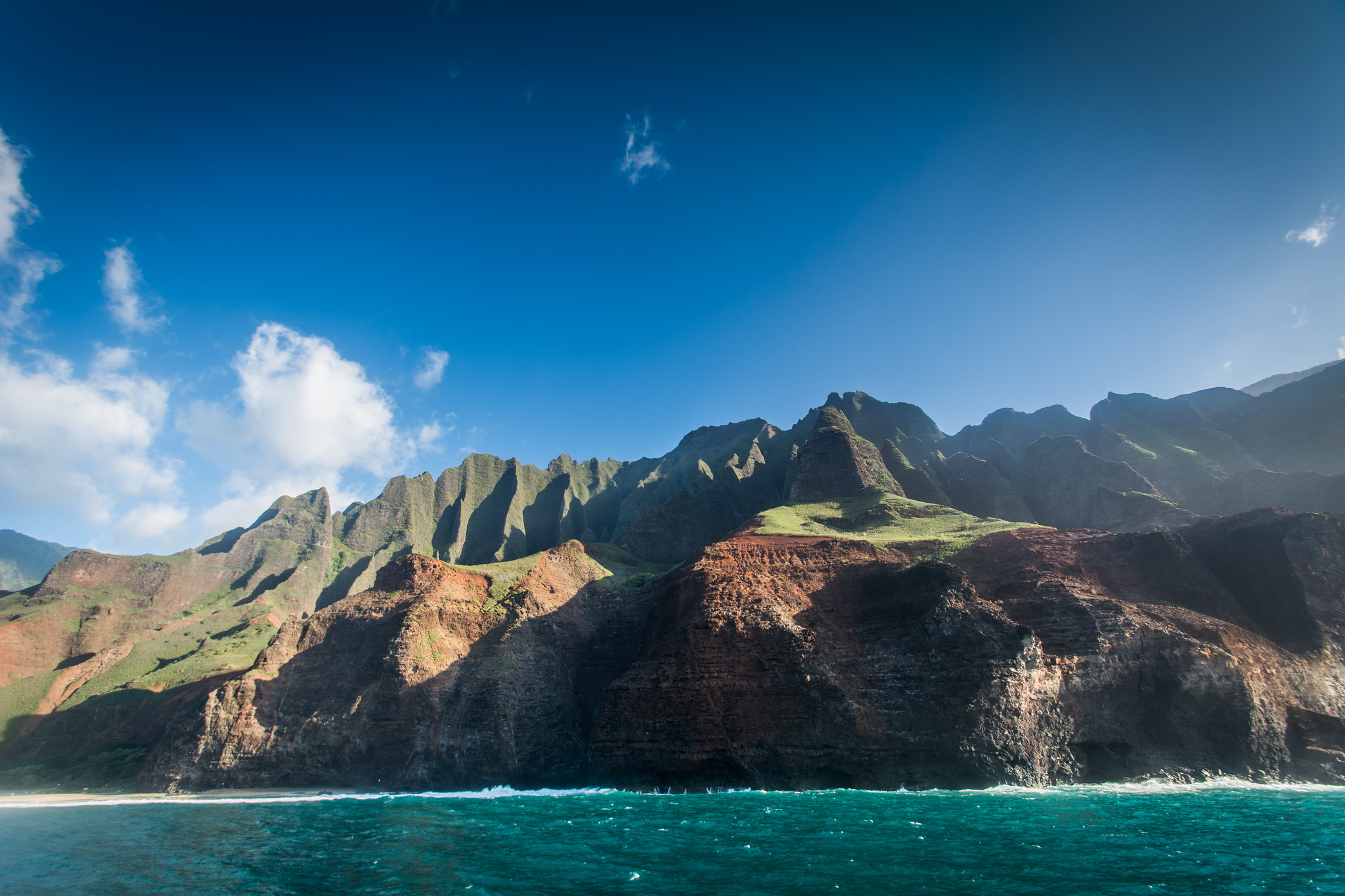 Island cliffside with clear blue water in the foreground and lightly cloudy blue sky in the background.