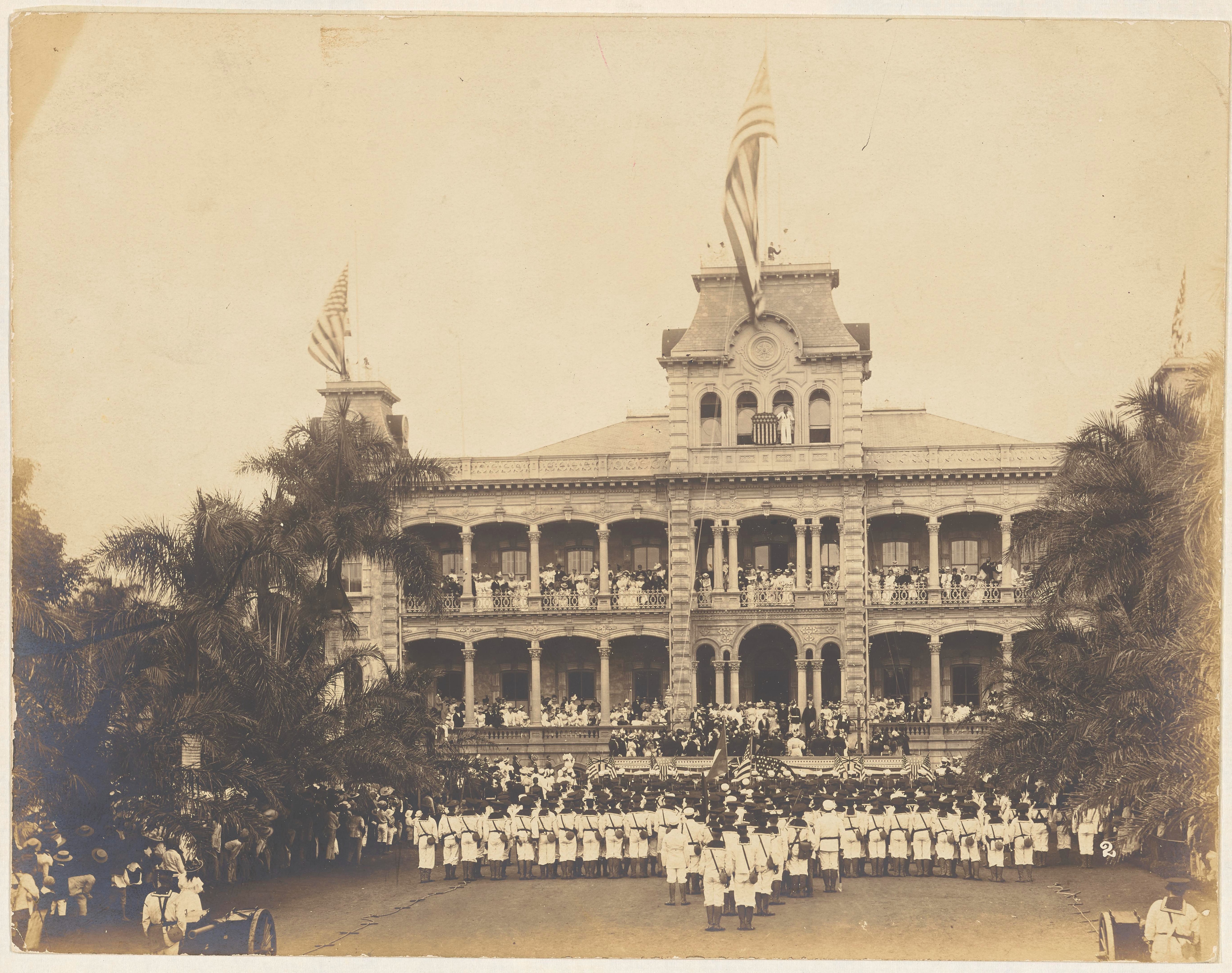 Large palatial building with three United States flags mounted on the roof. Military people stand facing the building in a ceremonial formation. Non-military people stand on the building's balcony facing the military people.