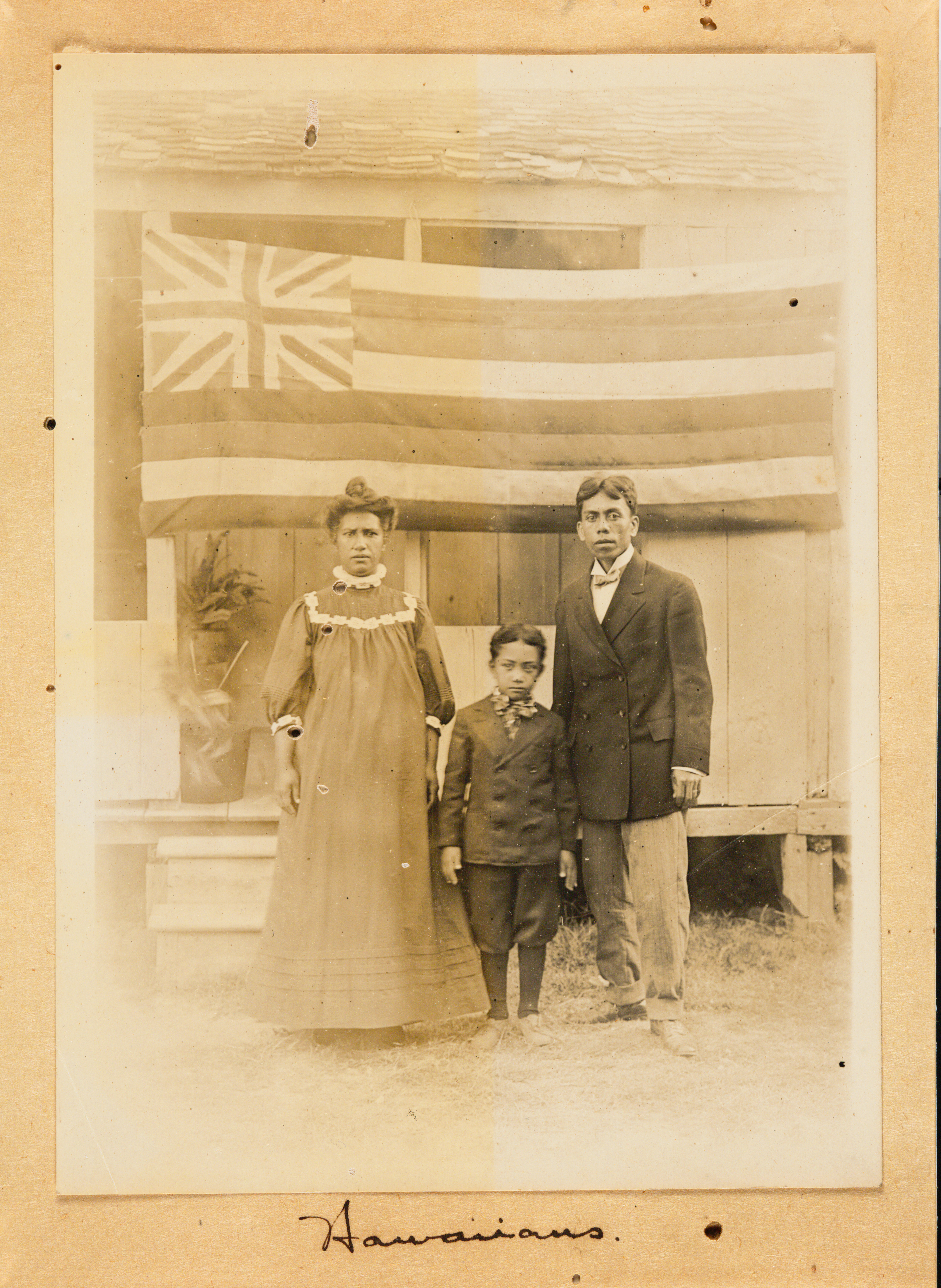 A Hawaiian man, woman, and boy pose for a photograph wearing Western clothing. Behind them hangs the flag of the Kingdom of Hawaii. Manuscript text below the image reads: Hawaiians.