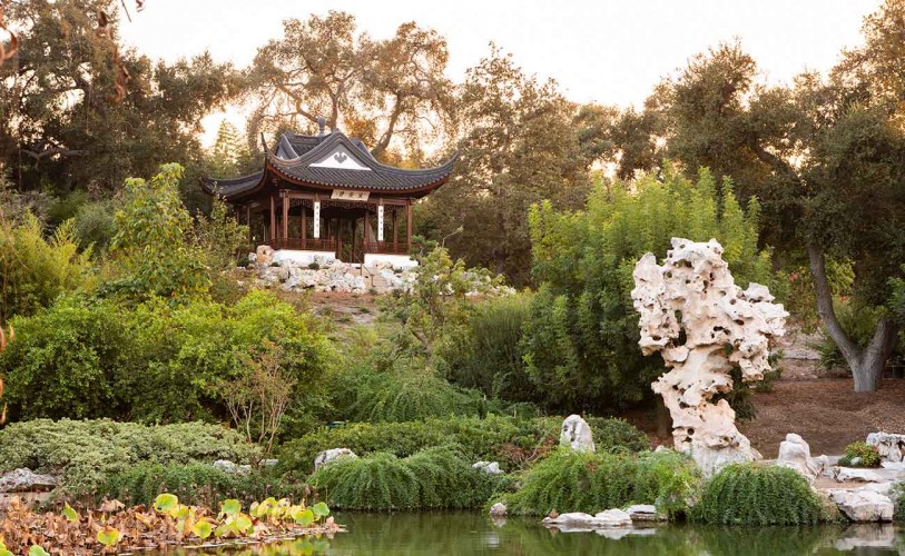 Garden with standing water in the foreground, a large rock next to the water ,and a one-room structure with a curved roof on a hill above the water.