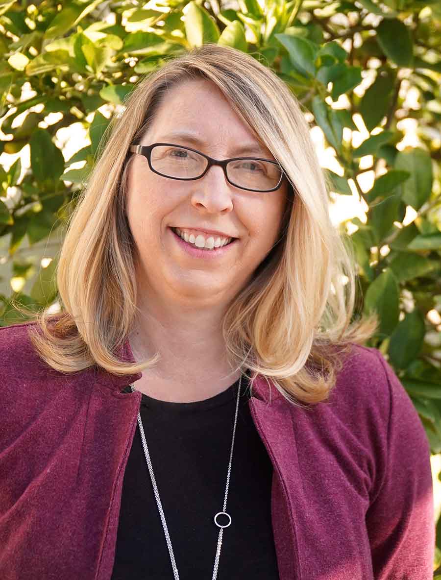 A white woman in a black shirt and red sweater smiles in front of a leafy background.