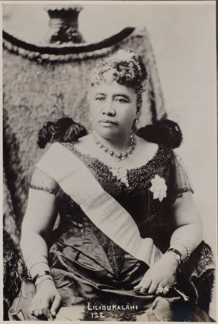 Hawaiian woman wearing a dress, cross-body sash, and jewelry poses for a photograph. Manuscript text at the bottom of the photograph reads: Lilioukalani. 12E.