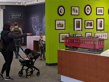 A woman pushes a stroller through a gallery that features a miniature replica of an old Pacific Electric trolley car.