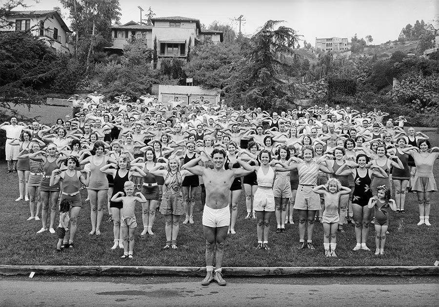 A group of 100 or so people in bathing suits and shorts, make a "crab" pose, showing off the muscles in their arms. 