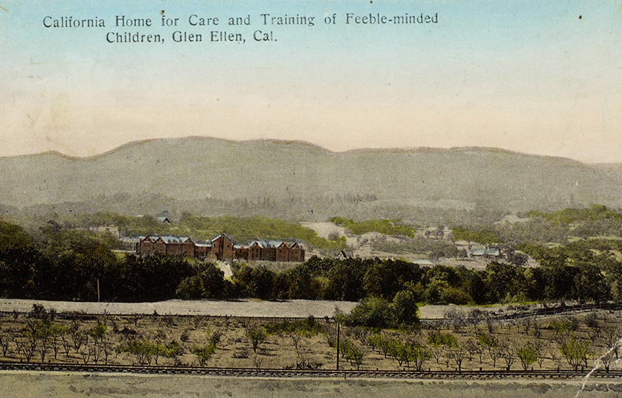 A postcard photograph of a large building among trees. A train track runs in the foreground.