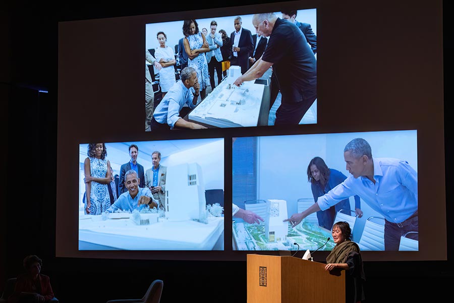 Billie Tsien stands at a podium with images of the Obamas behind her.