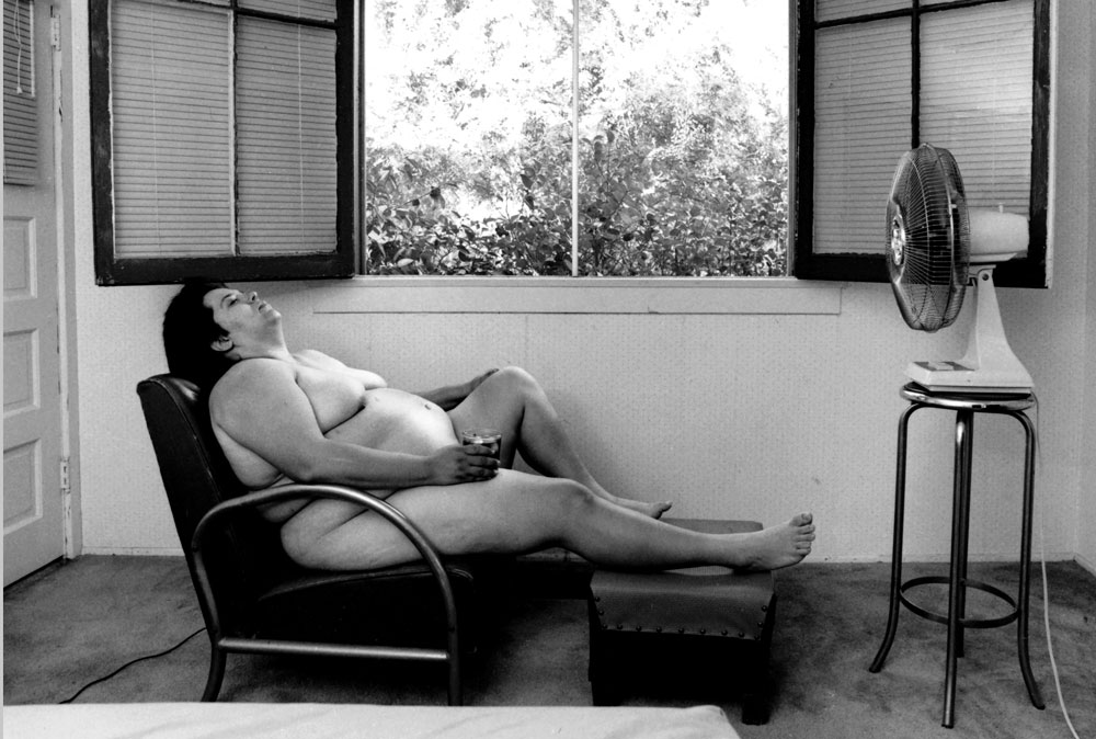 A nude person reclining in a chair by an open window with a fan on.