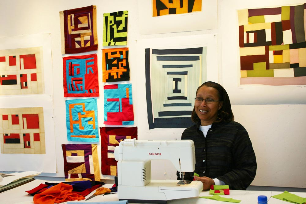 A person sits behind a sewing machine; the wall behind them is covered in small, colorful quilts.