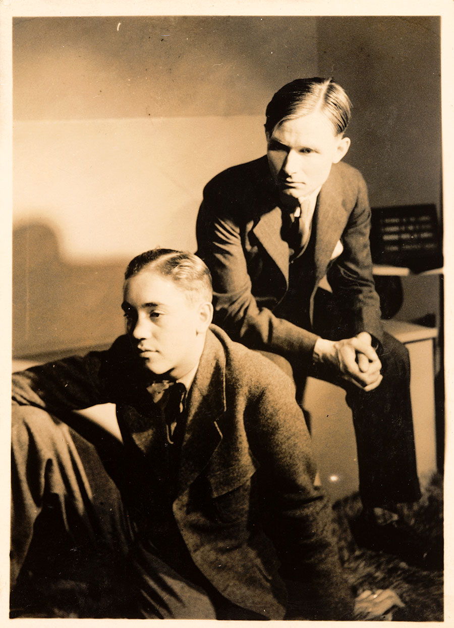 Black-and-white photo of two young men, one sitting on a bench, the other on the floor.