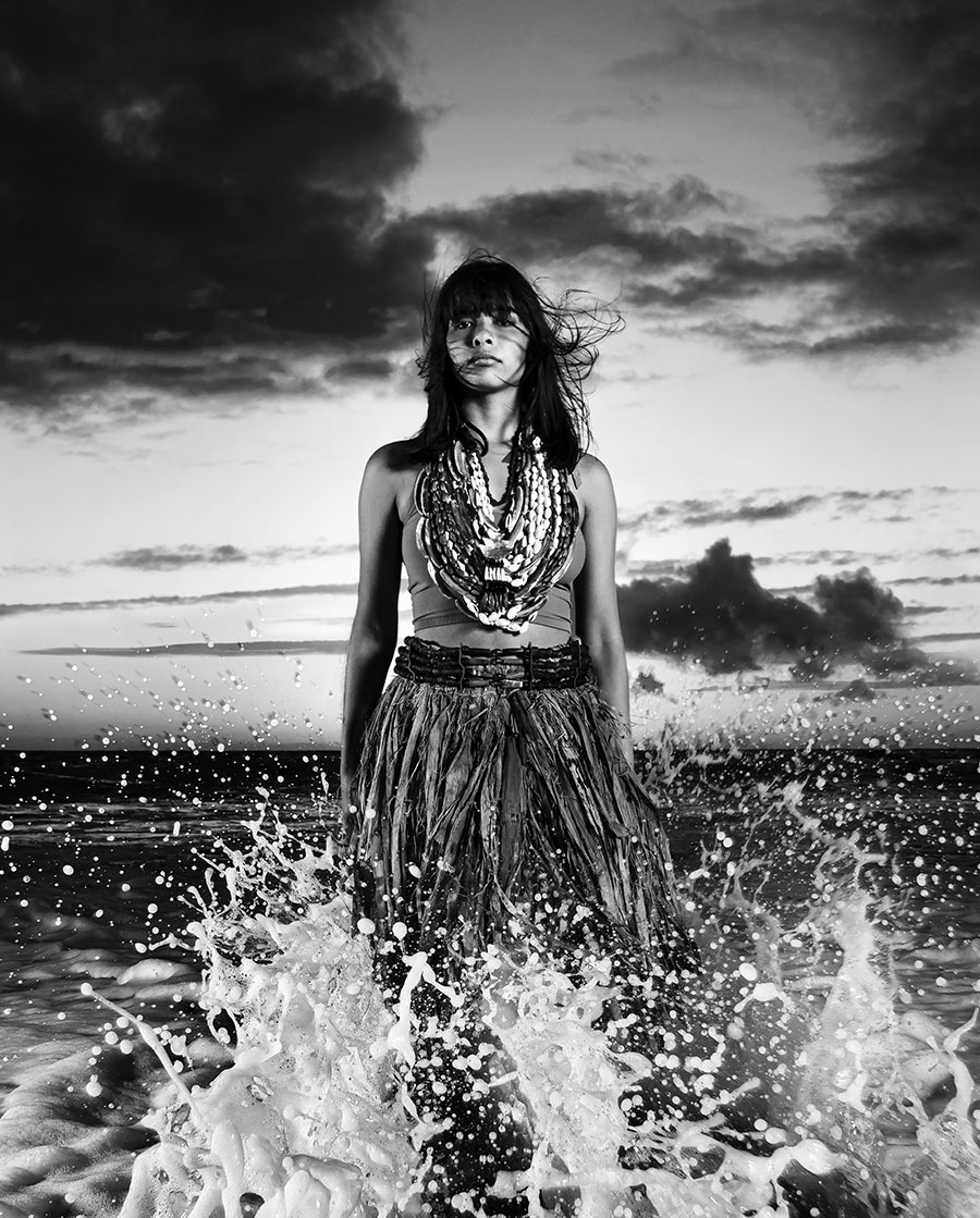 Black-and-white photo of a young adult dressed in multiple necklaces standing among splashing ocean waves.