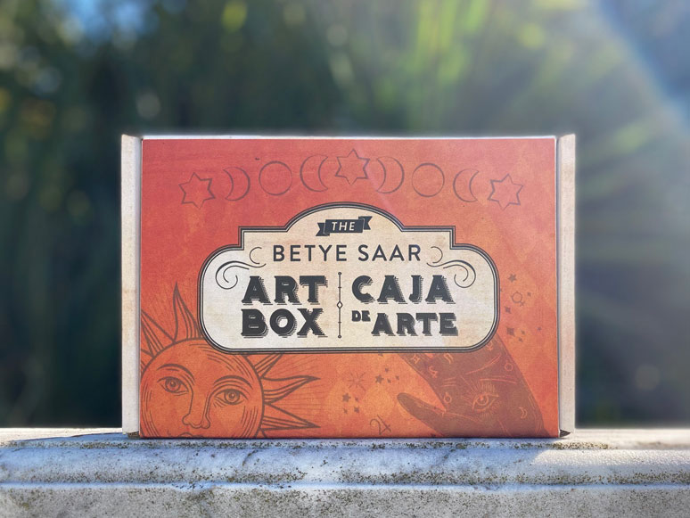 An orange box with celestial motifs, a label reads "The Betye Saar Art Box | Caja De Arte."