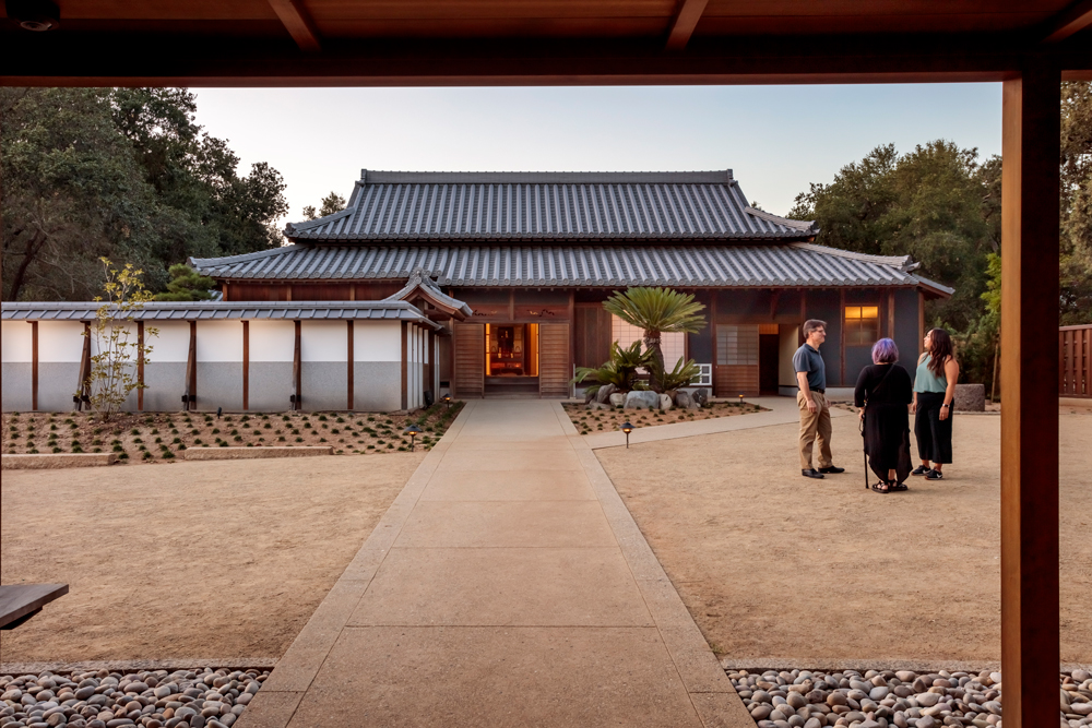 Three people stand in a gravel courtyard, near a path leading to a traditional Japanese house.