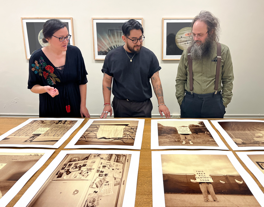 Three people stand over a table observing a series of photo prints.