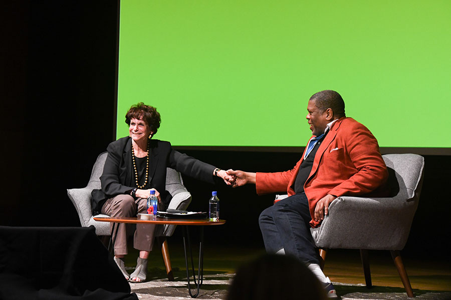 Two people on a stage, sitting next to each other and shaking hands.