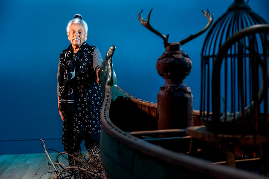 Artist Betye Saar stands near a wood canoe in a blue room.