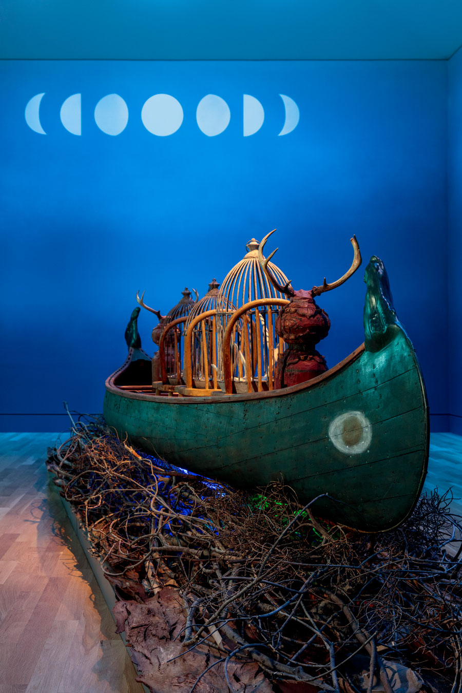 A wood canoe sits on top of a bed of dry branches in a blue room with phases of the moon painted on the wall.