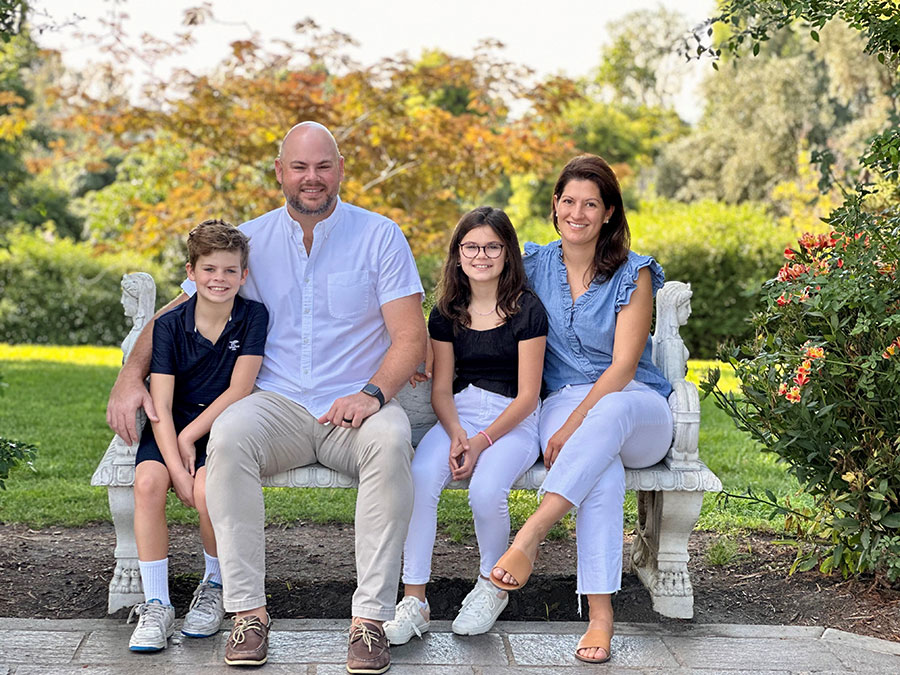 A family sits on a concrete bench in a garden, smiling for the camera.