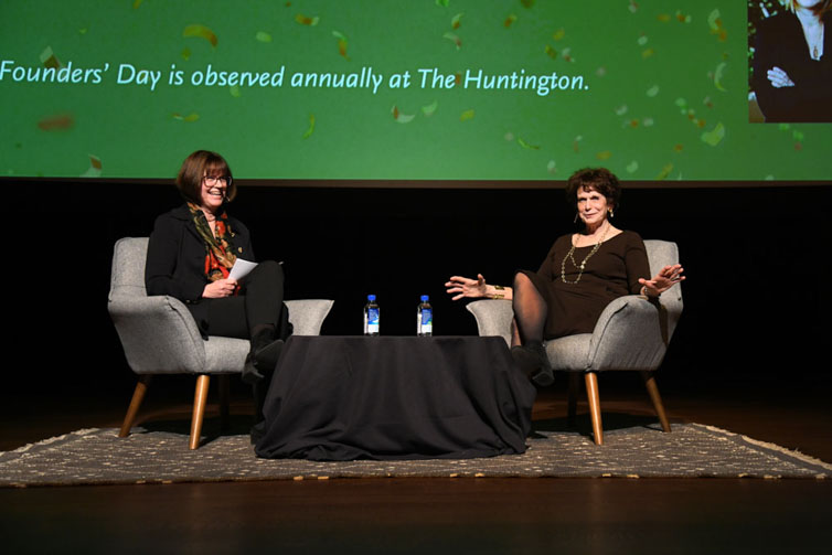 Two people sit in chairs on a stage in front of a large screen.