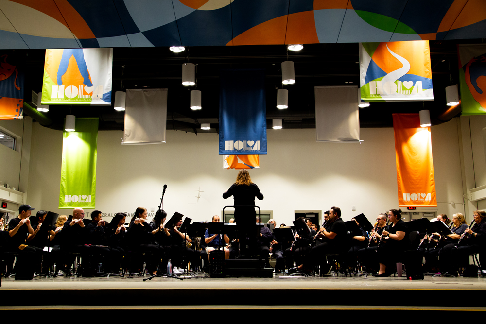A conductor stands in front of an orchestra on a stage with colorful banners.